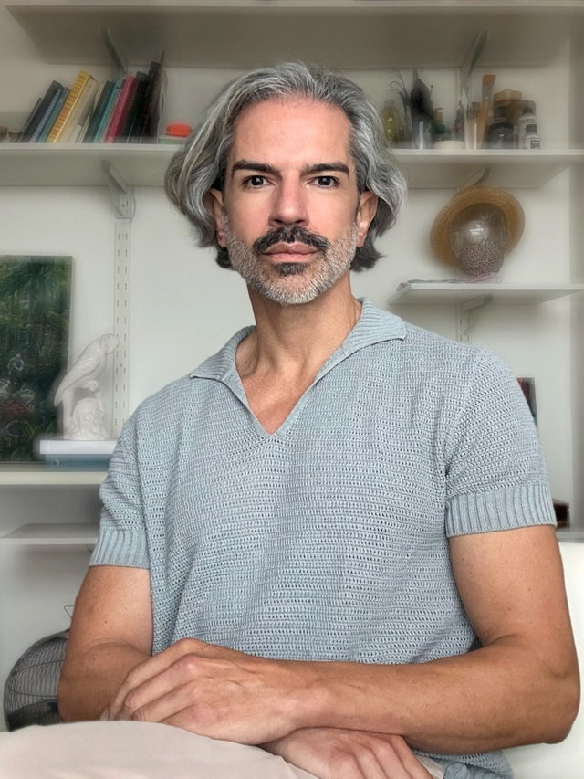 Homem de cabelo grisalho e camisola de malha cinzenta, sentado em frente a prateleiras com livros, pincéis e um papagaio de cerâmica.
